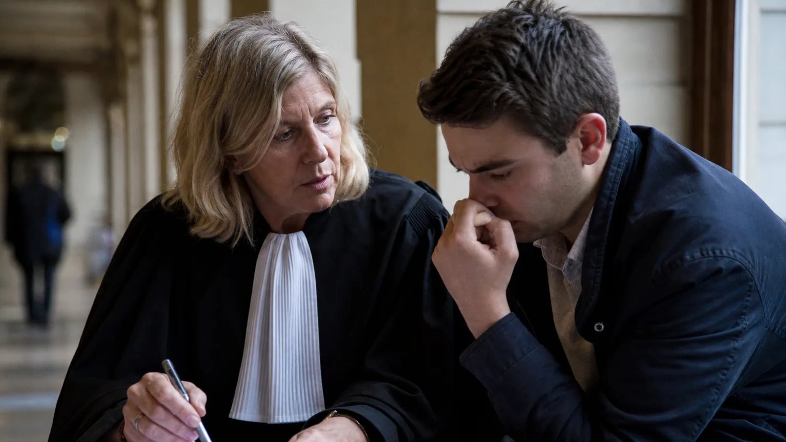 Un avocat en robe noire conseille un client inquiet dans le couloir d'un tribunal.