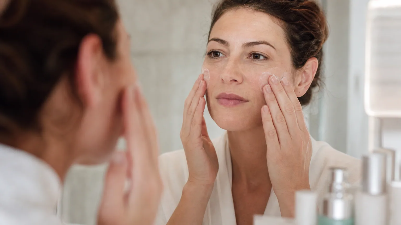 Une femme applique une crème hydratante sur son visage devant un miroir le matin