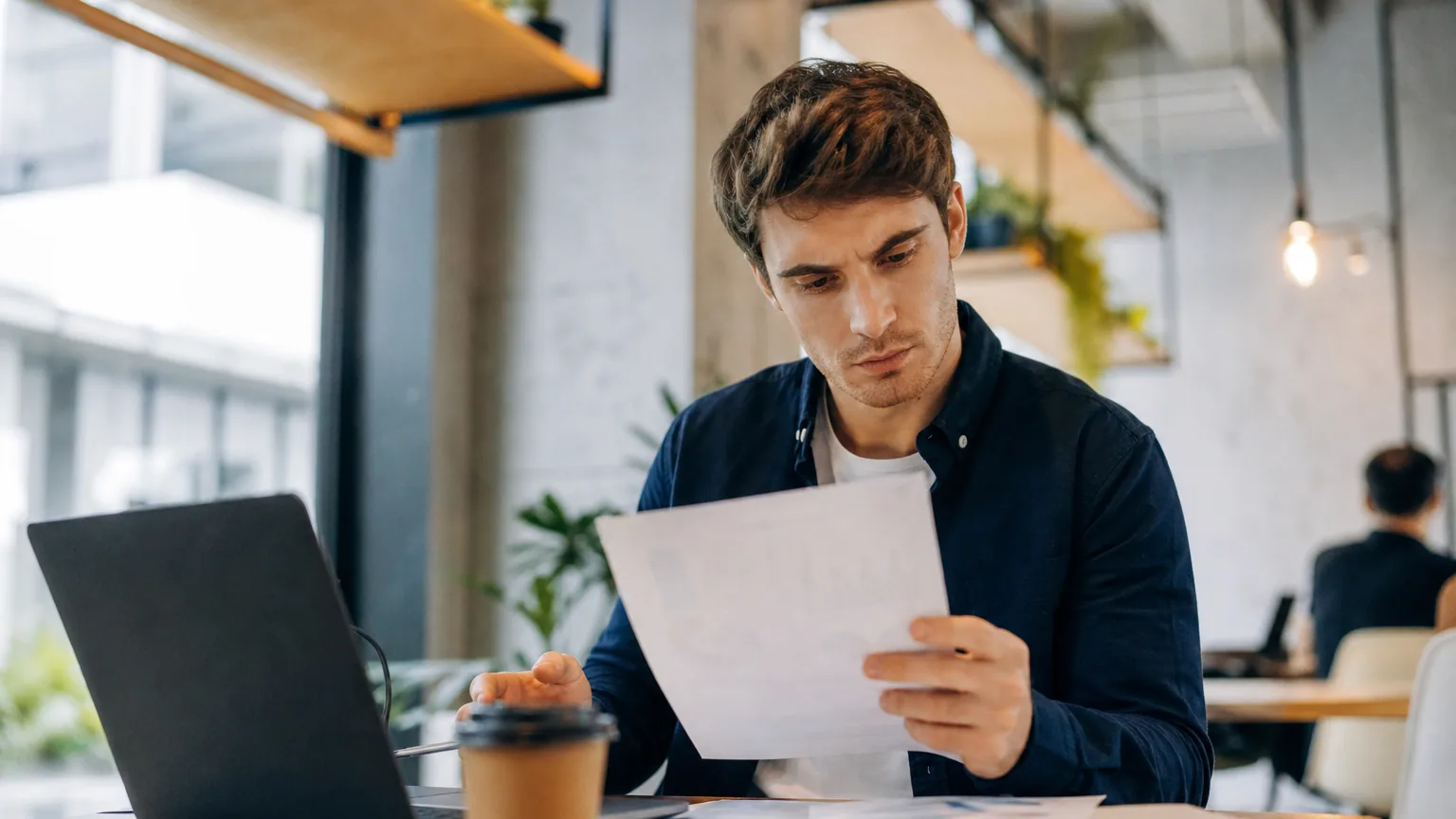 Jeune entrepreneur comparant des documents et un ordinateur portable à son bureau, l'air concentré