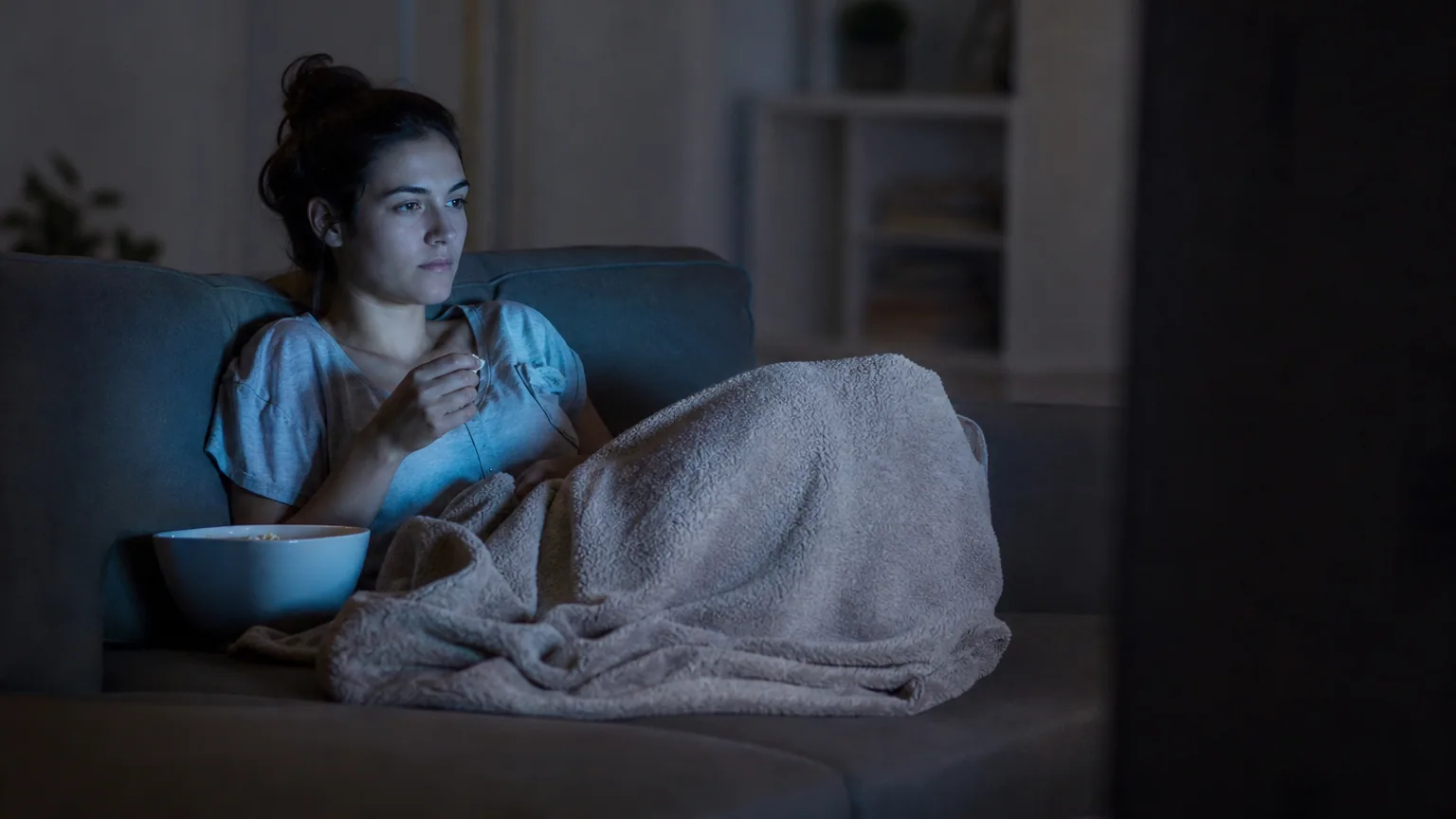 Une femme regarde une série tard le soir, seule sur un canapé éclairé par la lumière de la télévision