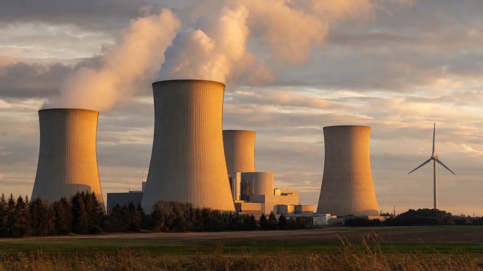 Centrale industrielle émettant de la vapeur au coucher du soleil, avec une éolienne en arrière-plan