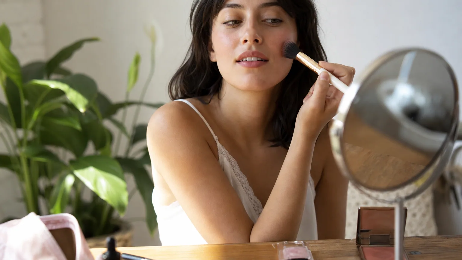 Jeune femme appliquant du blush au pinceau devant un miroir avec quelques produits de maquillage